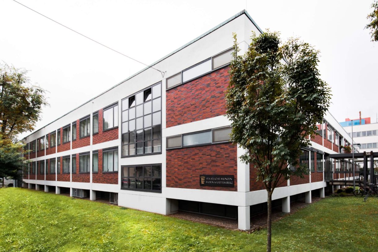 Gebäude der Staatlichen Münzen am Standort Stuttgart, mit roter Klinker-Fassade, weißen Trennelementen, Bäumen und einem Grünstreifen auf der Anlage